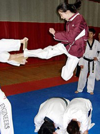 Una mujer salta y se cierne unos cinco pies en el aire sobre dos personas inclinadas en el suelo. La mujer tiene el pie derecho extendido en una patada de karate, rompiendo una tabla.